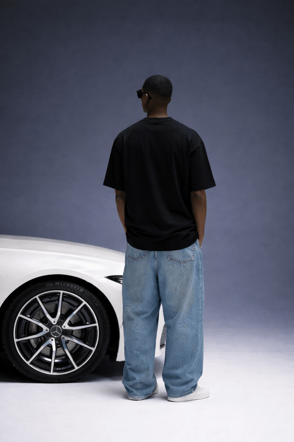 Person wearing a black t-shirt and blue jeans standing next to a white car against a gray background